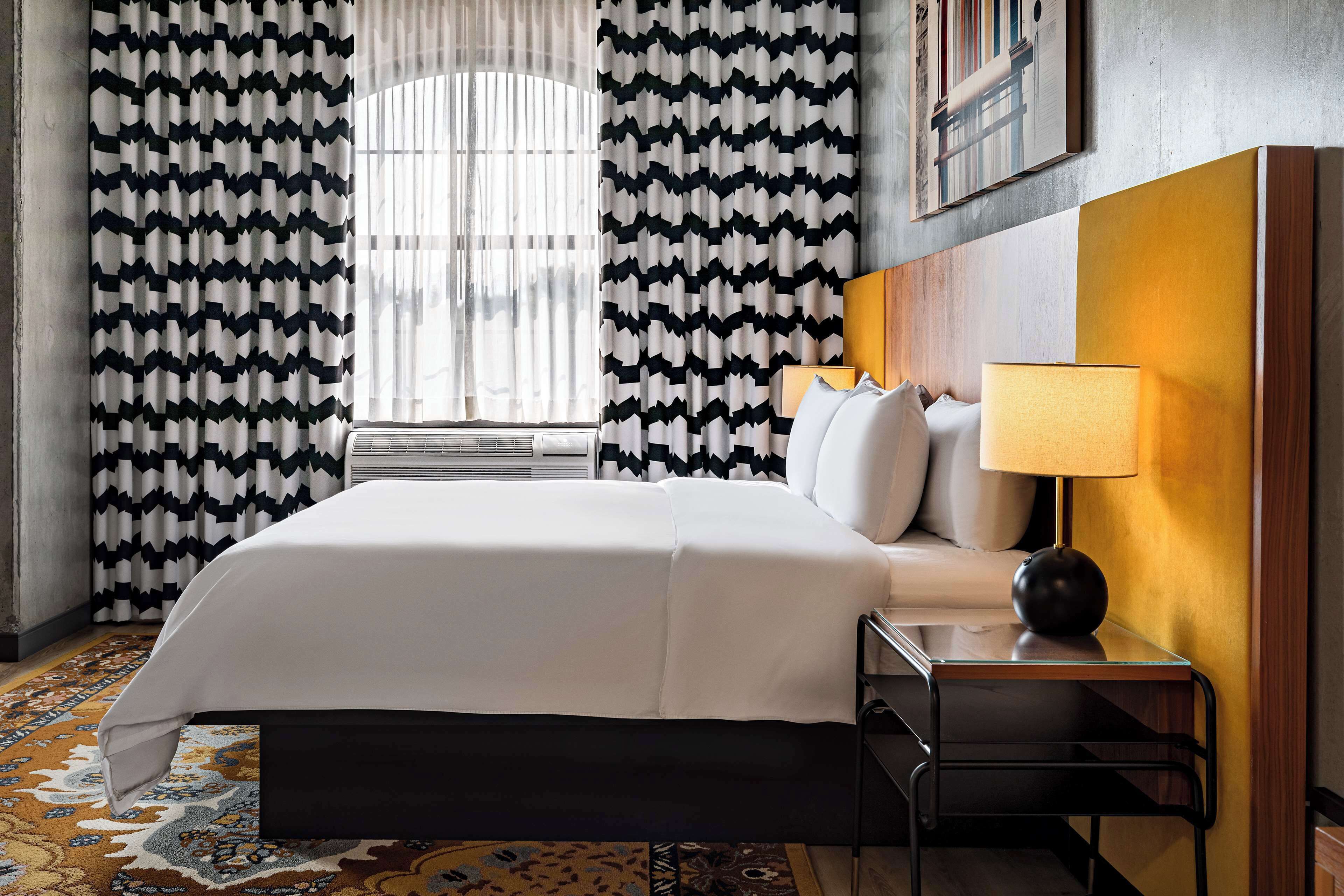 Ein Hotelzimmer mit Bett, Nachttisch, Lampe und gemusterten Vorhängen.