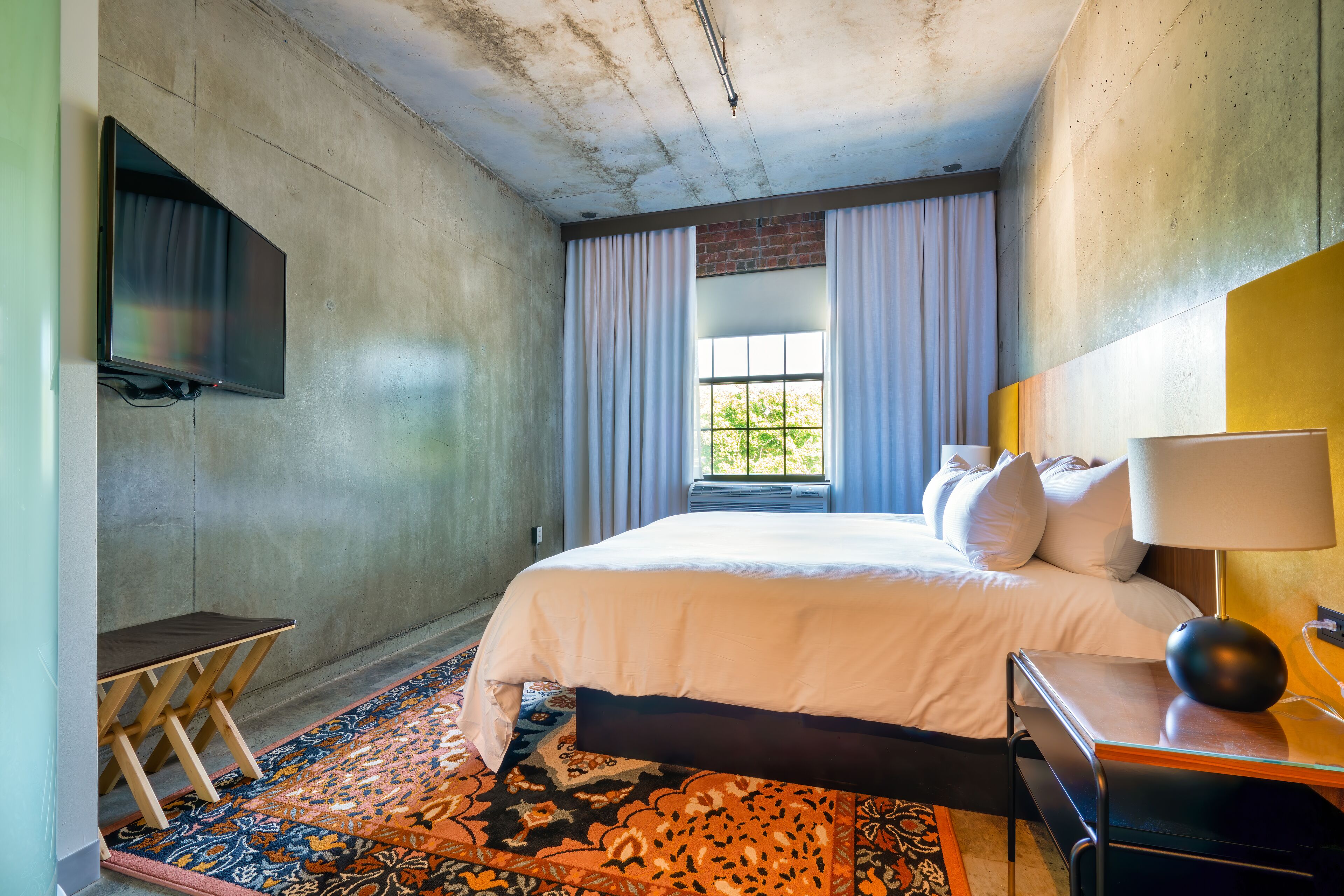 A hotel room with a large bed, a flat-screen TV mounted on the wall, a small table, and a window with curtains.