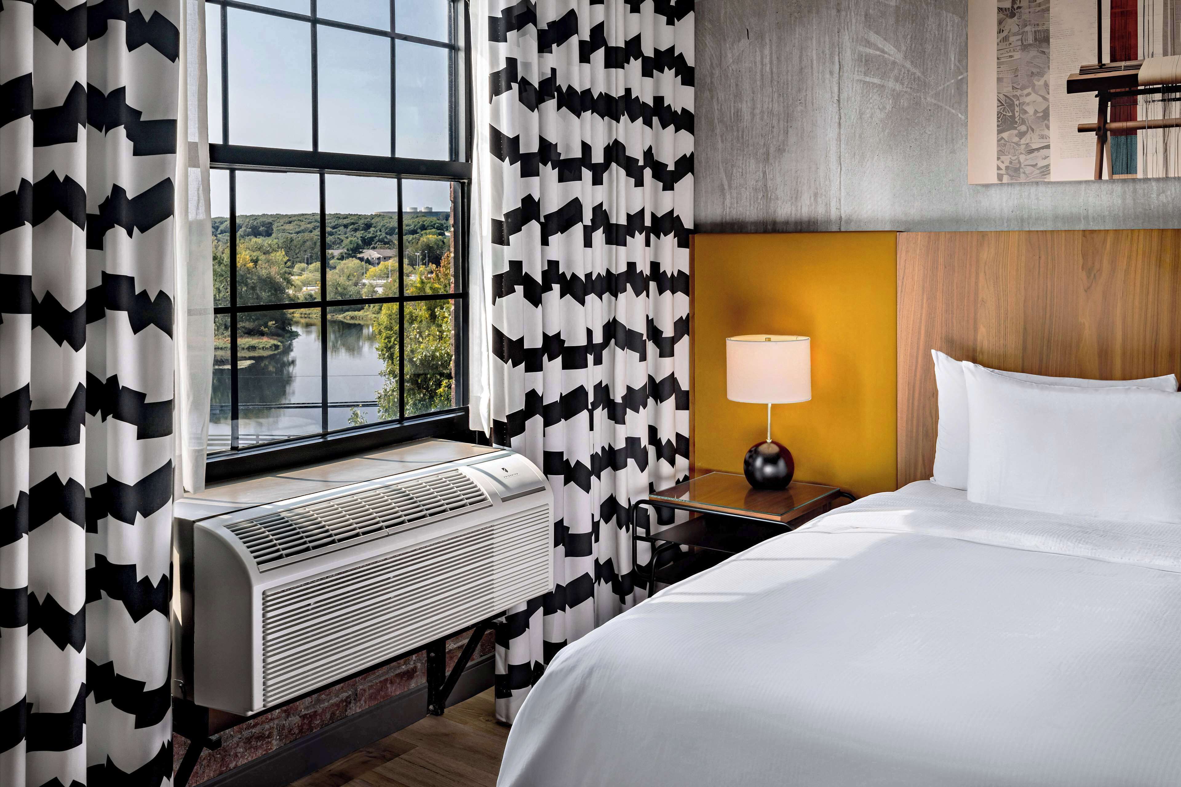 A hotel room with a large window, a bed, a nightstand with a lamp, and a wall-mounted air conditioner.