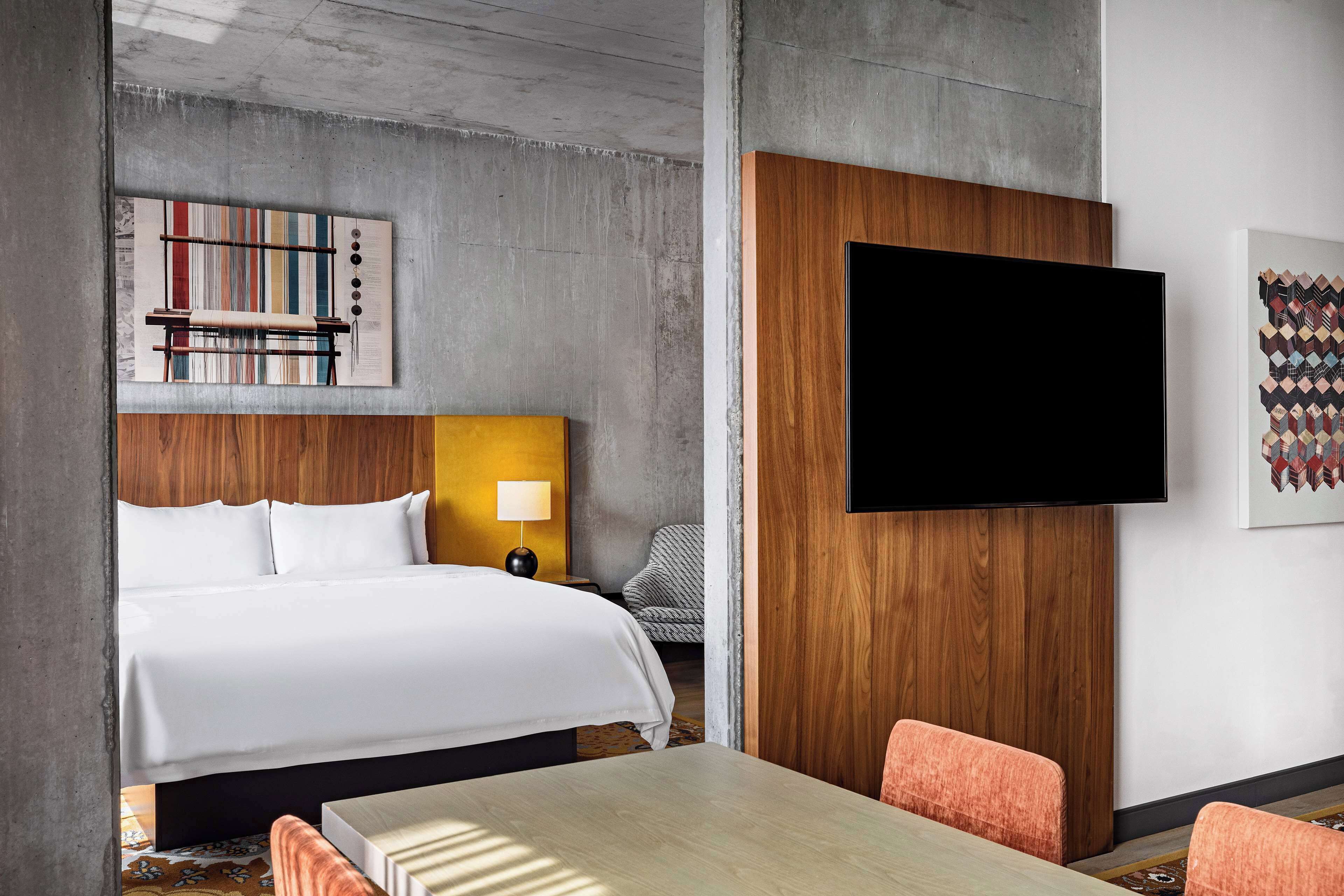Ein Hotelzimmer mit einem großen Bett, einem flachbildfernseher an der Wand, einem hölzernen Kopfteil, einem kleinen Tisch und zwei orangefarbenen Stühlen.