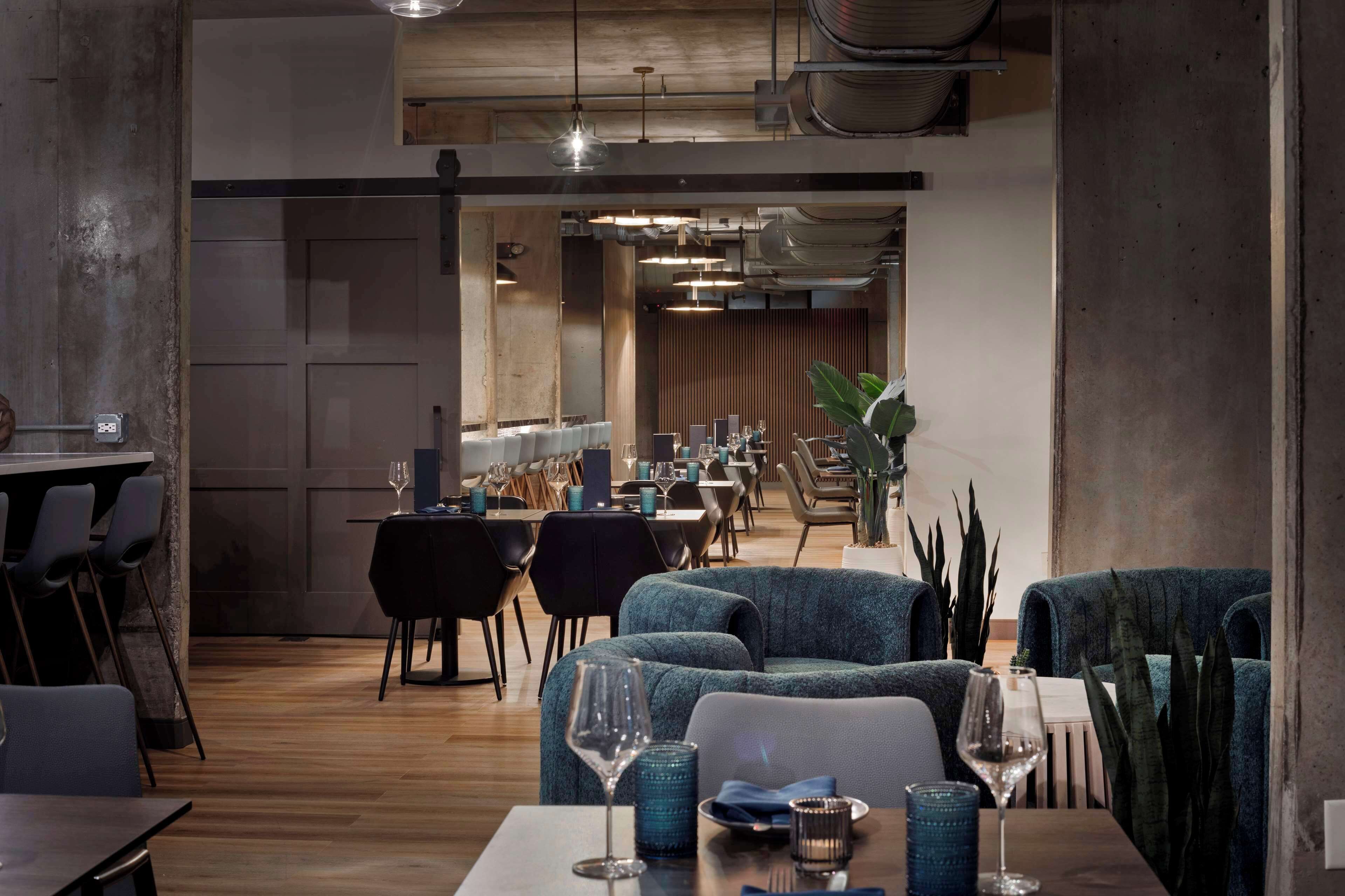 A modern restaurant interior with wooden flooring, blue upholstered seating, and a long dining area with tables and chairs.
