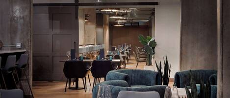 A modern restaurant interior with wooden flooring, blue upholstered seating, and a long dining area with tables and chairs.