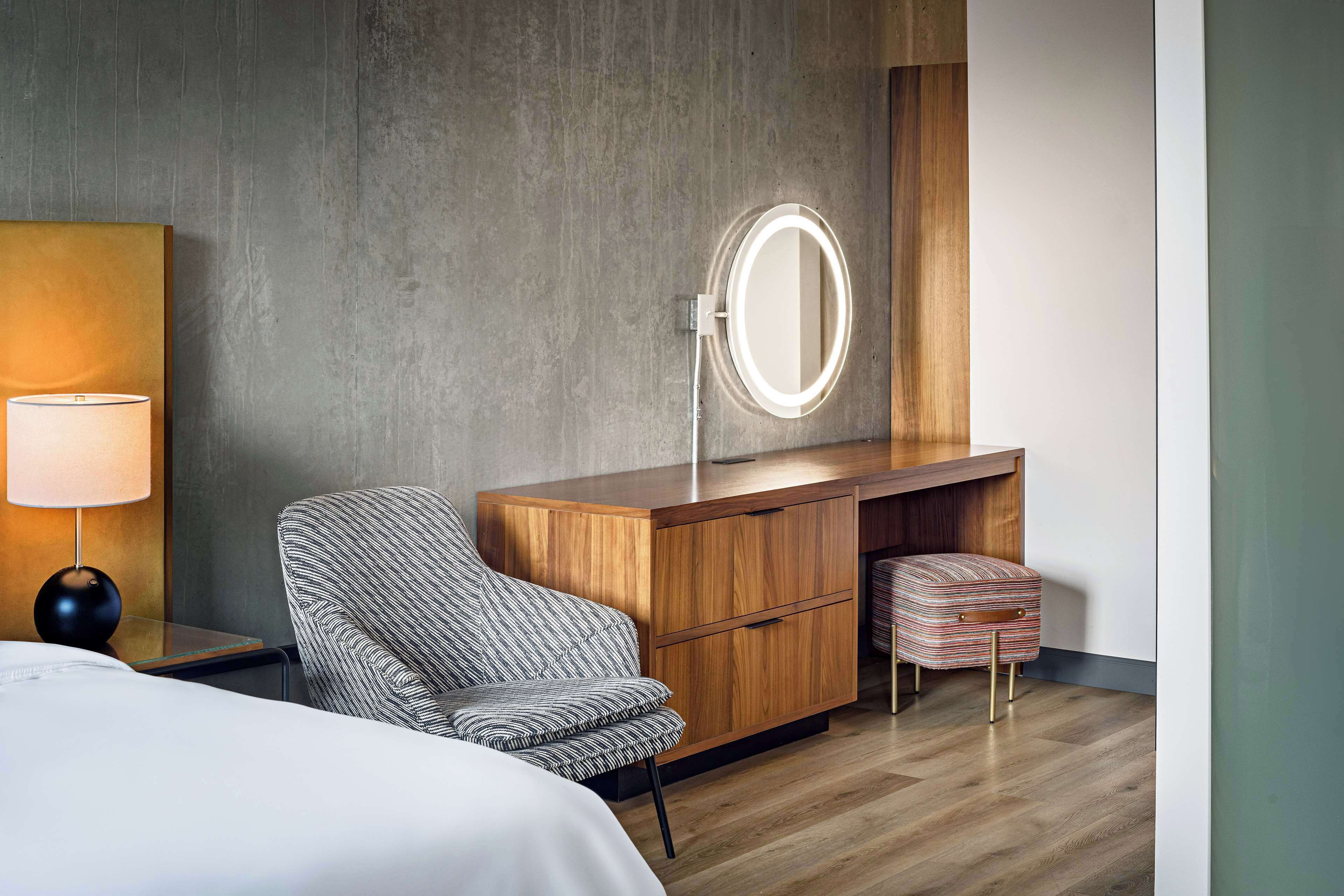 Una habitación de hotel moderna con un tocador de madera, un espejo redondo, un sillón a rayas, un pequeño reposapiés y una mesita de noche con lámpara.