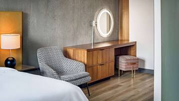 Una habitación de hotel moderna con un tocador de madera, un espejo redondo, un sillón a rayas, un pequeño reposapiés y una mesita de noche con lámpara.