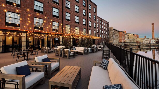 An outdoor seating area with sofas and tables, a brick building with large windows, and a river view. - The Loom Hotel, Tapestry Collection by Hilton (Warwick)