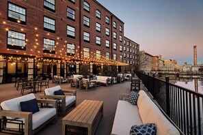 An outdoor seating area with sofas and tables, a brick building with large windows, and a river view. - The Loom Hotel, Tapestry Collection by Hilton (Warwick)