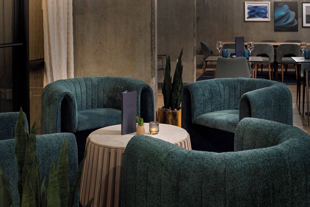 A modern lounge area with teal upholstered chairs, a round wooden table, and a potted plant.