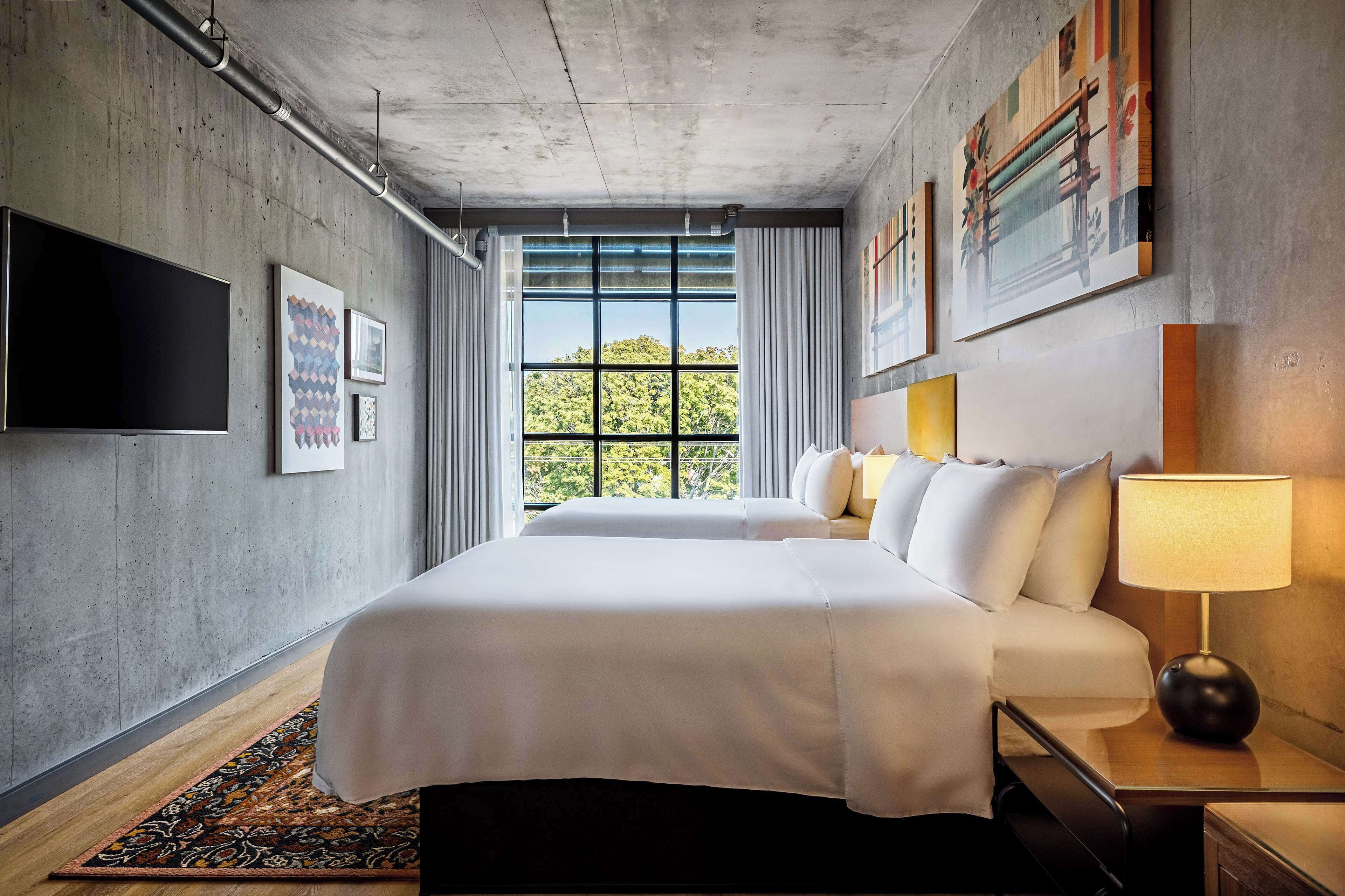 A modern hotel room with a large bed, a bedside table with a lamp, a TV mounted on the wall, and a window with a view of greenery.