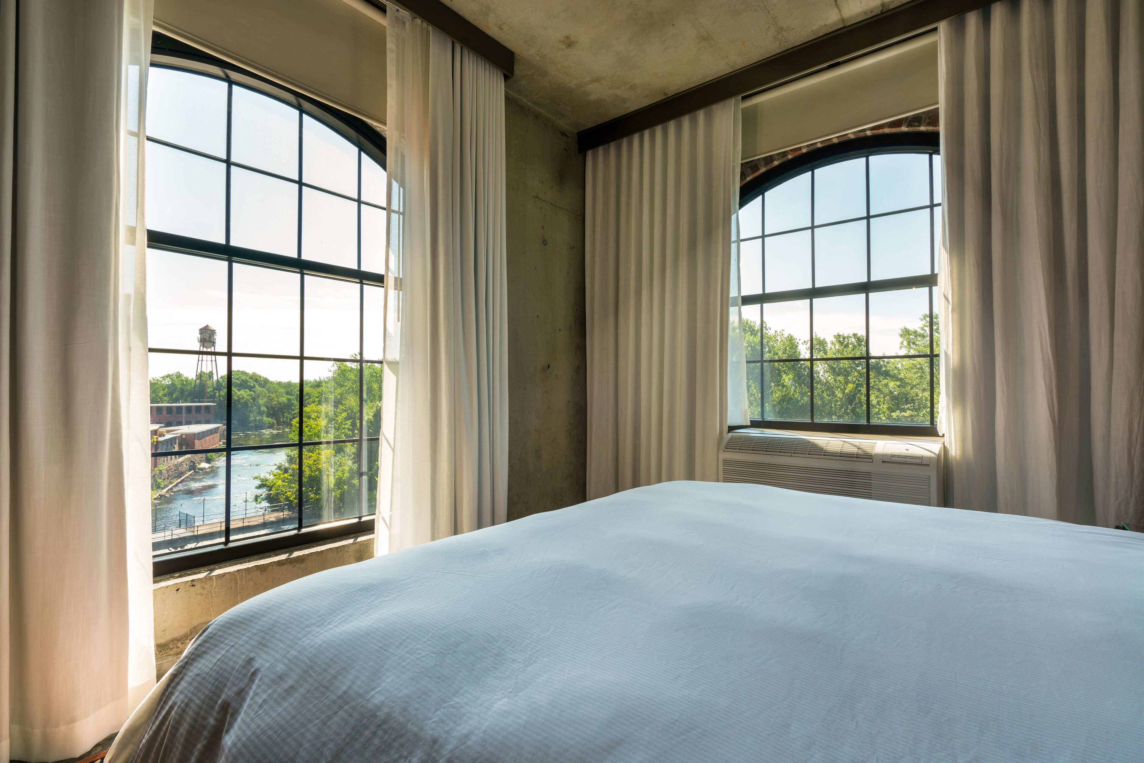 Hotelkamer met een groot bed, twee ramen met gordijnen en uitzicht op een rivier en groen.