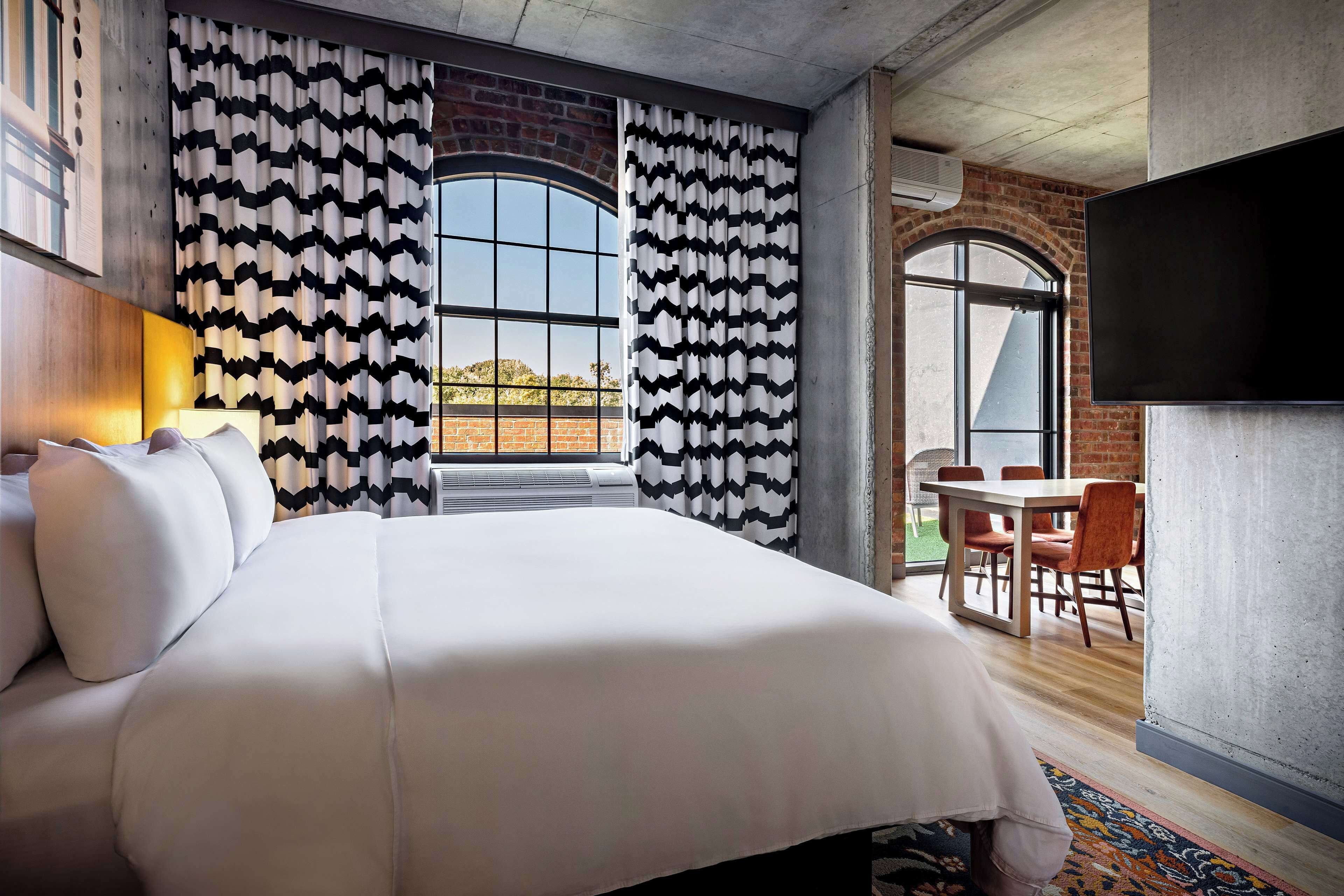 Habitación de hotel con una cama grande, un área de comedor con pared de ladrillo y un televisor montado en la pared.