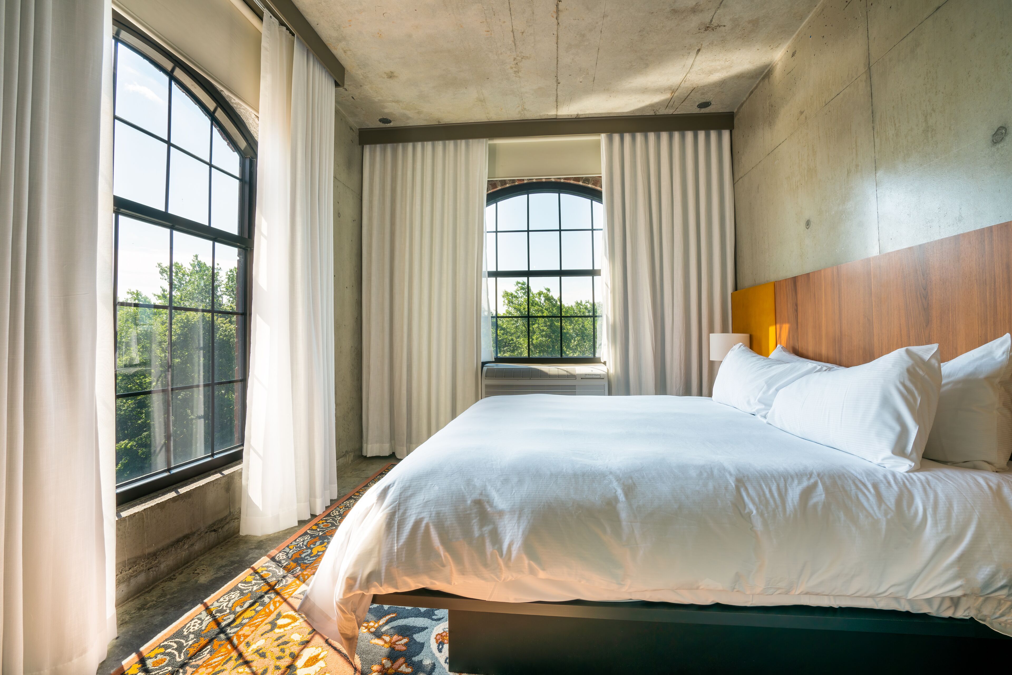 A large bed with white linens, a wooden headboard, and a view of greenery through a window.