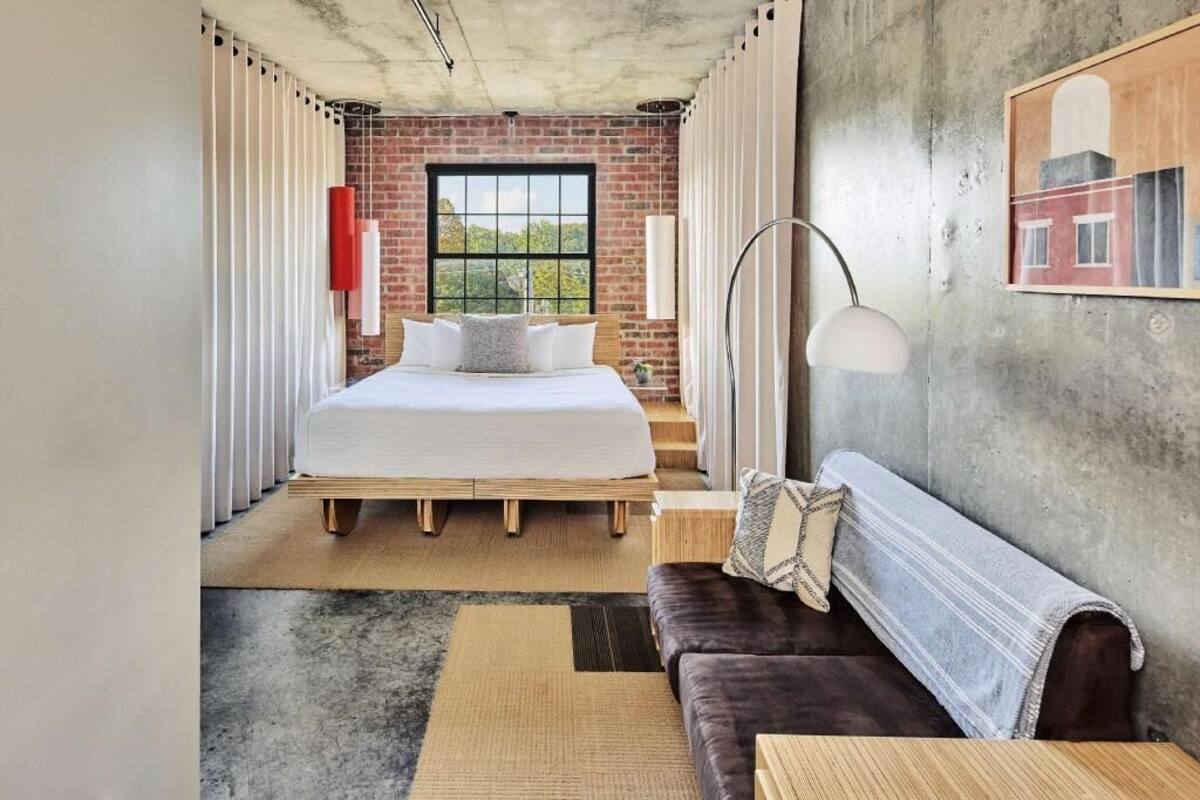 A modern bedroom with a brick wall, a large window, a bed, a sofa, and a lamp.