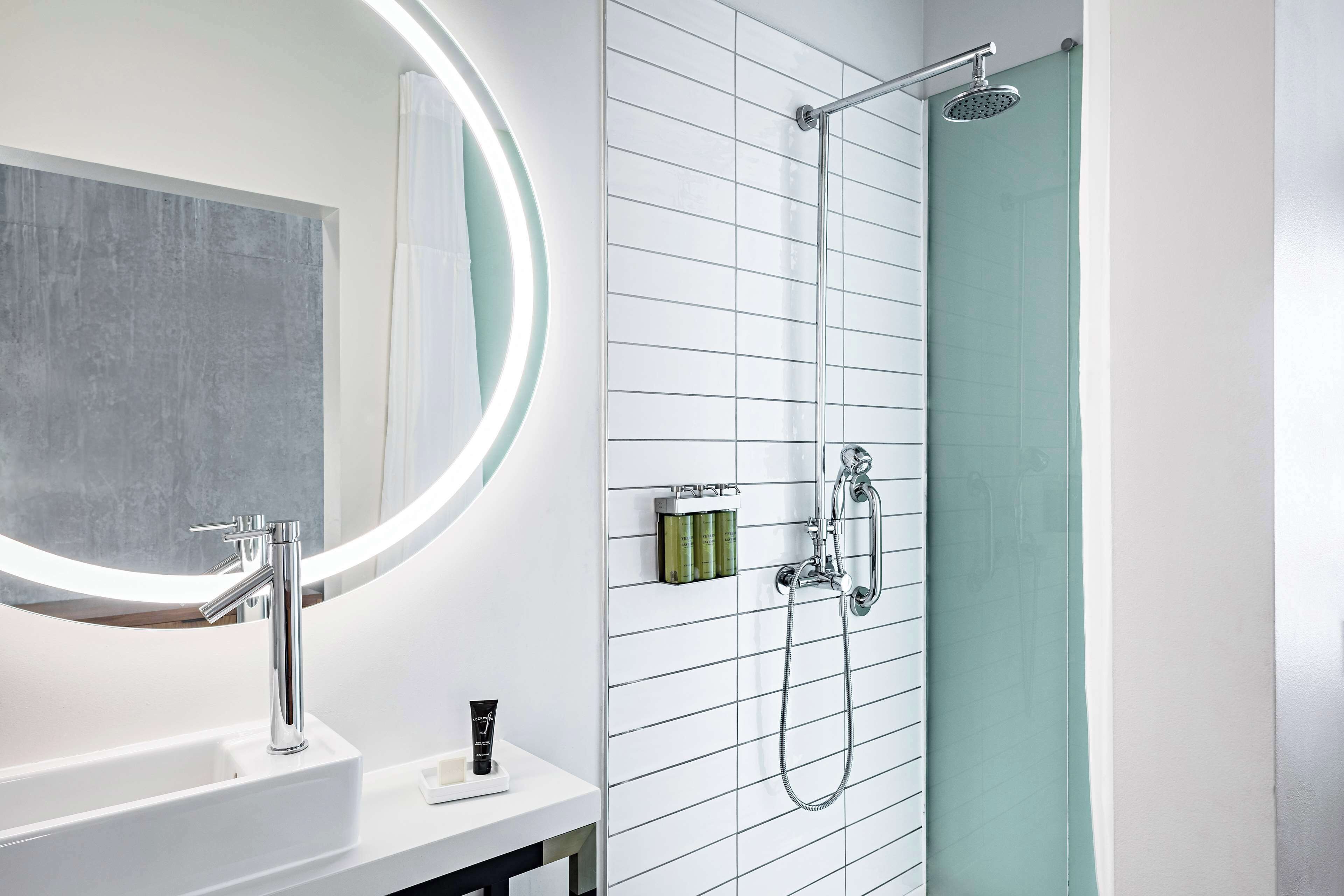 A modern bathroom with a white sink, a large mirror with a circular light, a shower with a handheld showerhead, and a green-tiled shower enclosure.