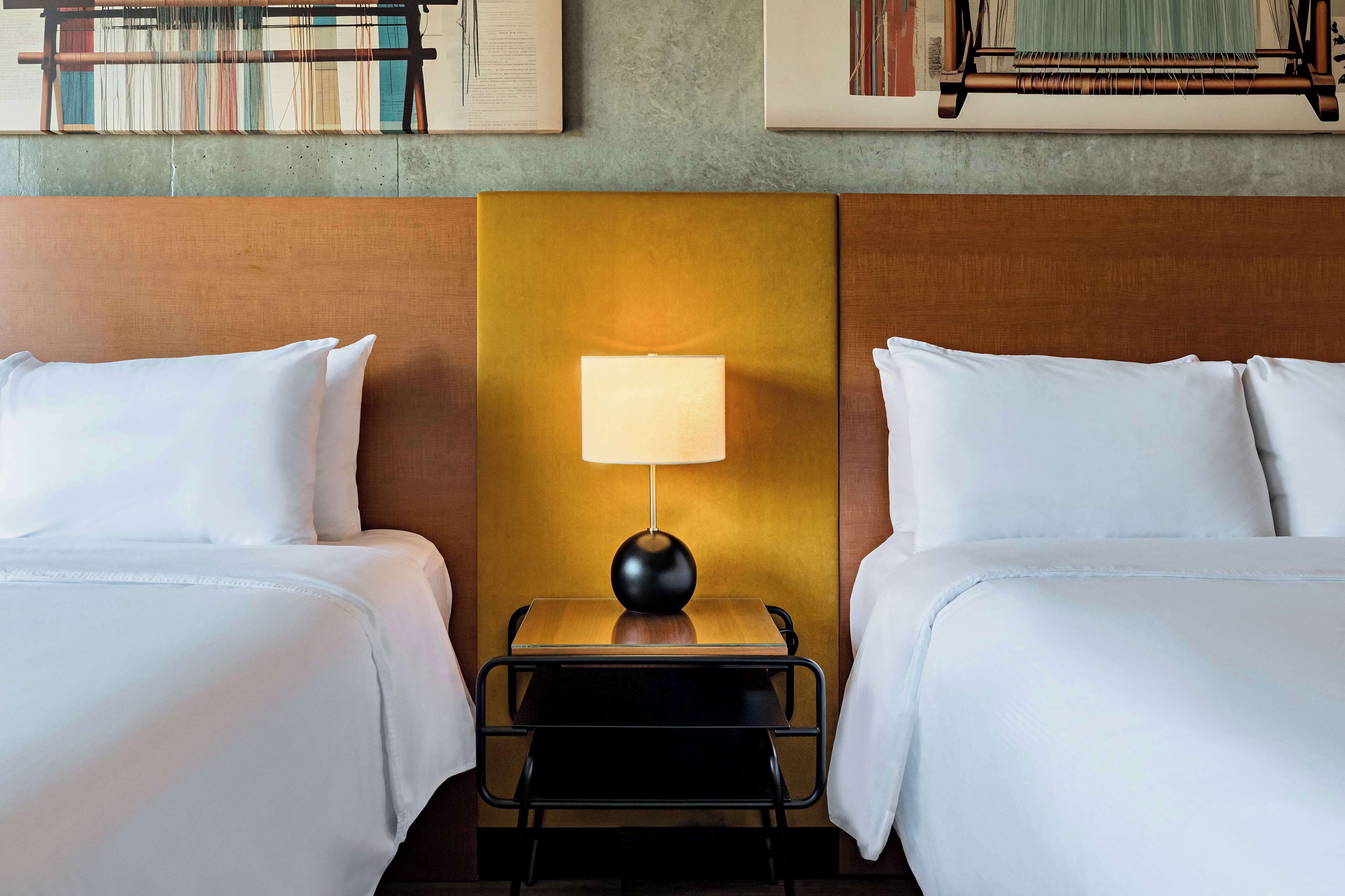 A hotel room with two beds, a nightstand with a lamp, and a wall-mounted bookshelf.
