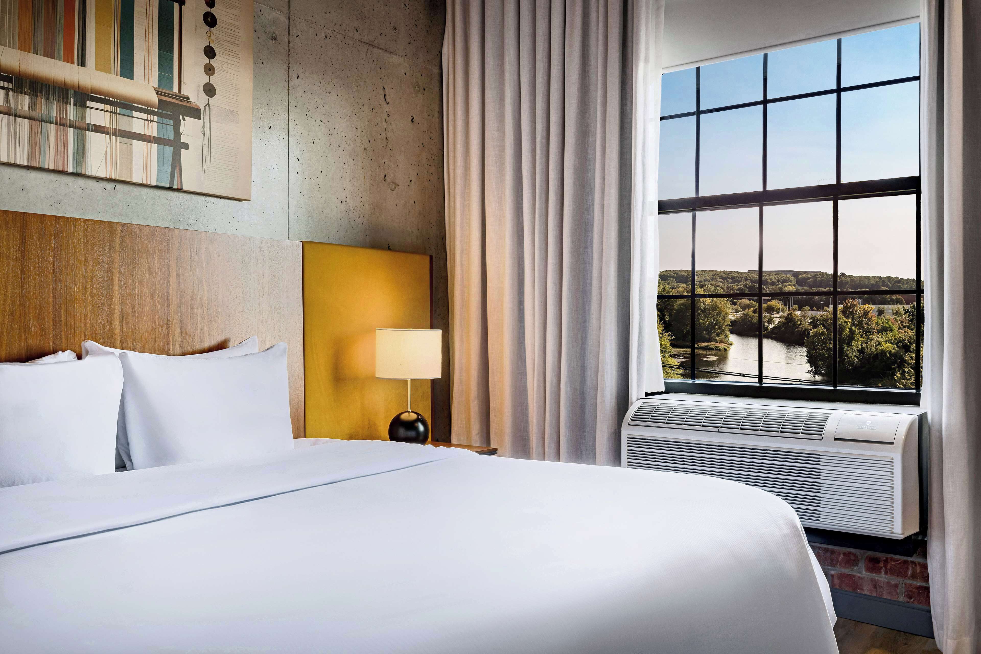 Habitación de hotel con una cama grande, una mesita de noche con lámpara, una ventana con vista a un río y árboles, y un equipo de aire acondicionado.