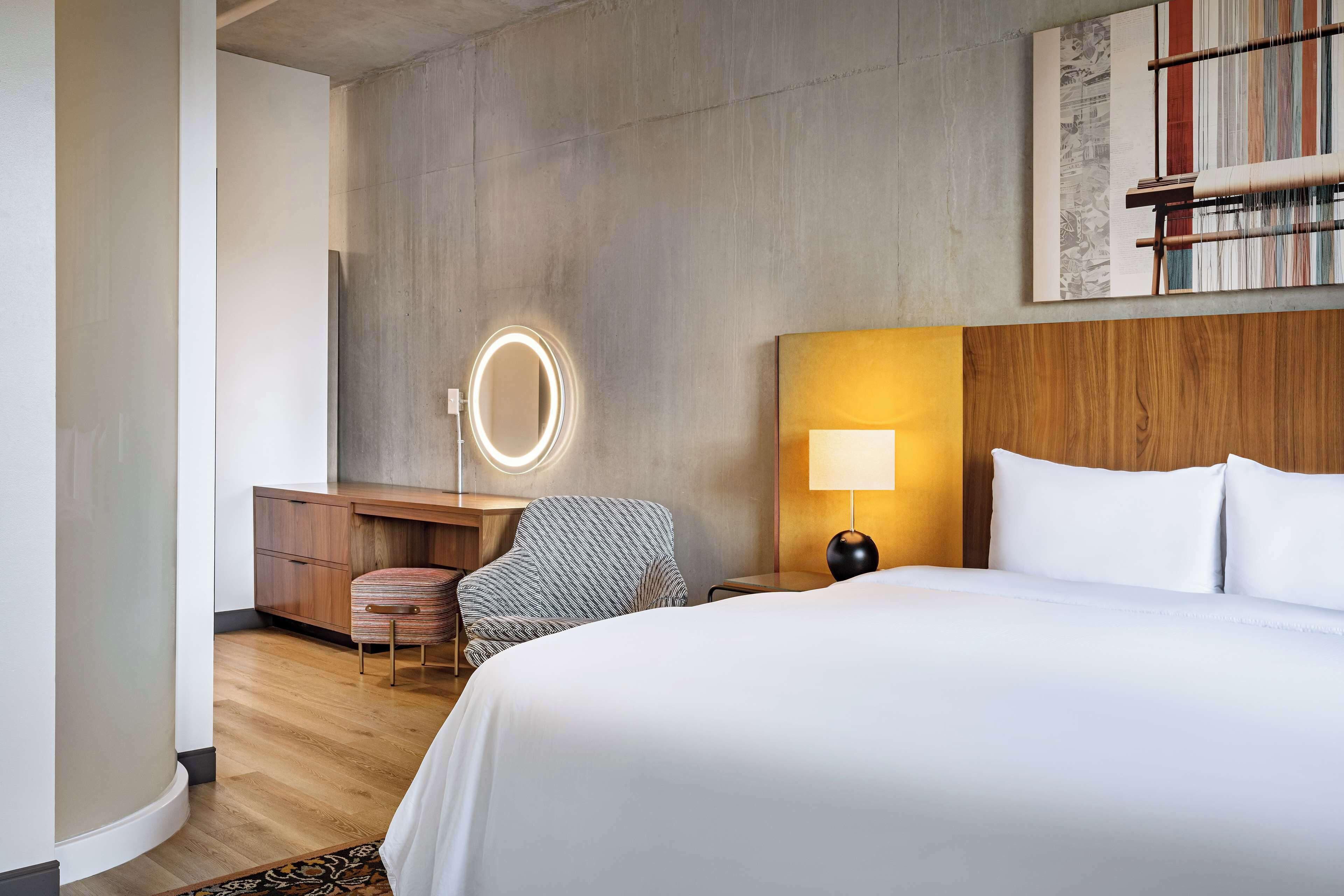 A hotel room with a large bed, a desk with a chair, a round mirror, a lamp, and a painting on the wall.