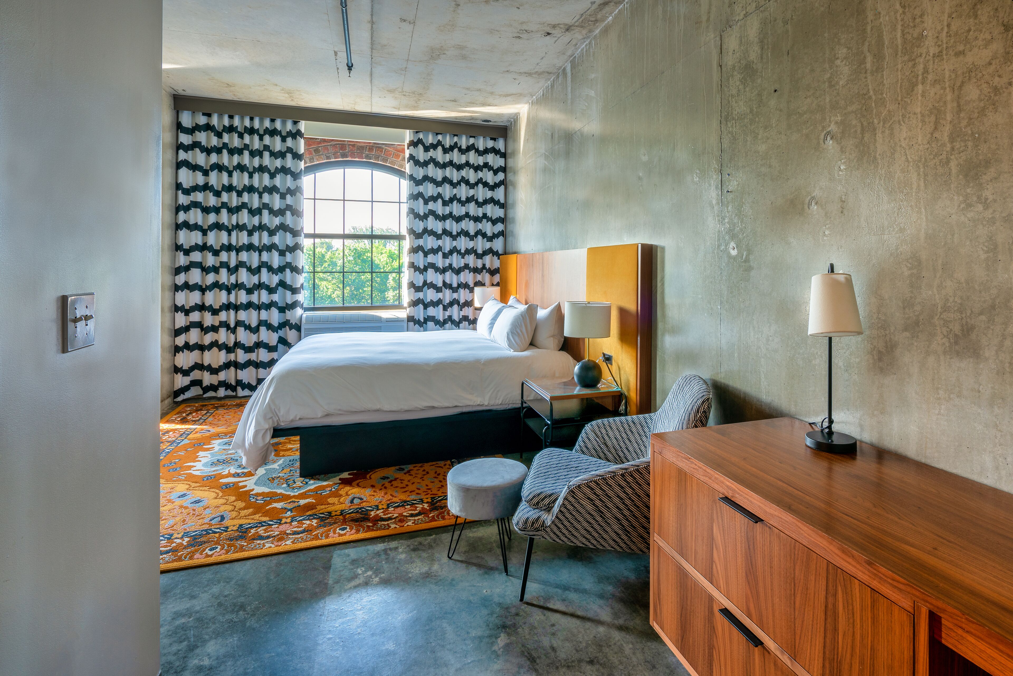 A bedroom with a bed, a nightstand, a chair, a wooden dresser, and a window with patterned curtains.