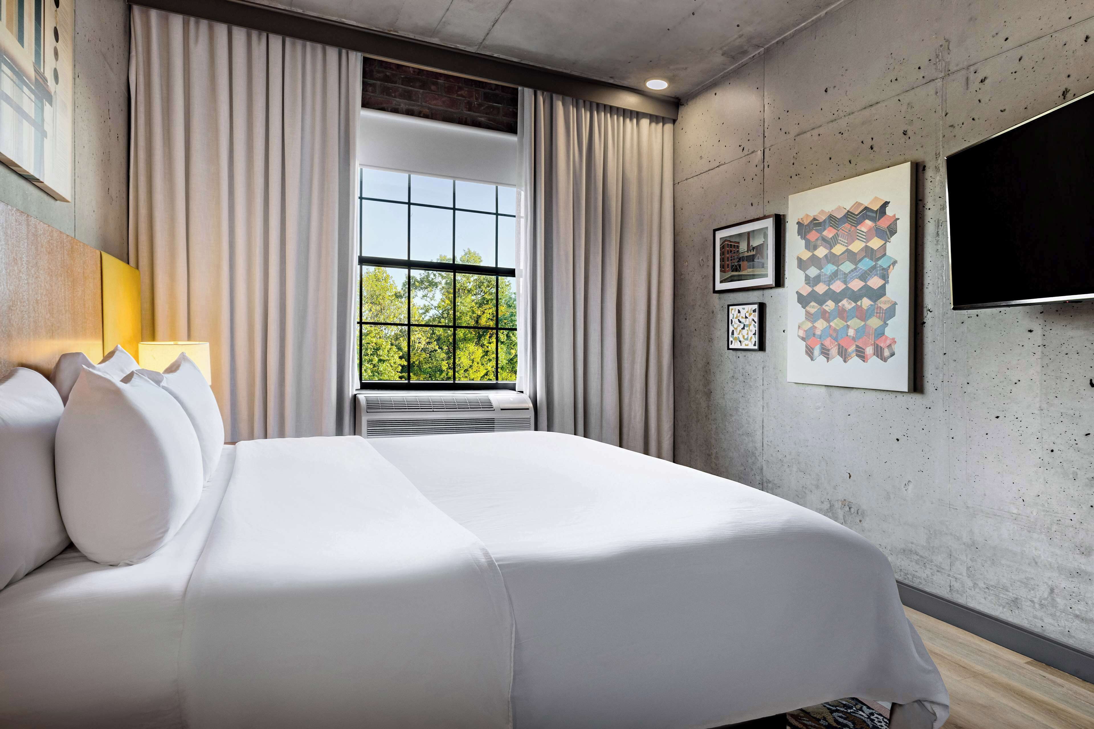 A modern hotel room with a large bed, a window with curtains, and artwork on the walls.