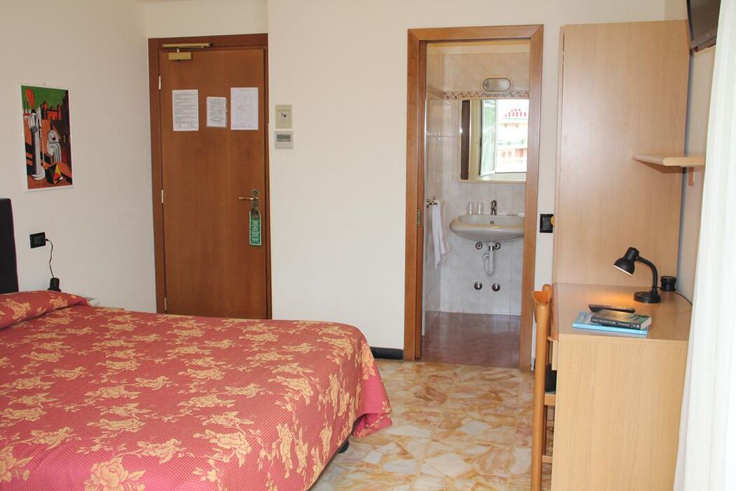 Ein Schlafzimmer mit Bett, einer Holztür, einem Badezimmer mit Waschbecken und Spiegel sowie einem Schreibtisch mit Lampe und Büchern.