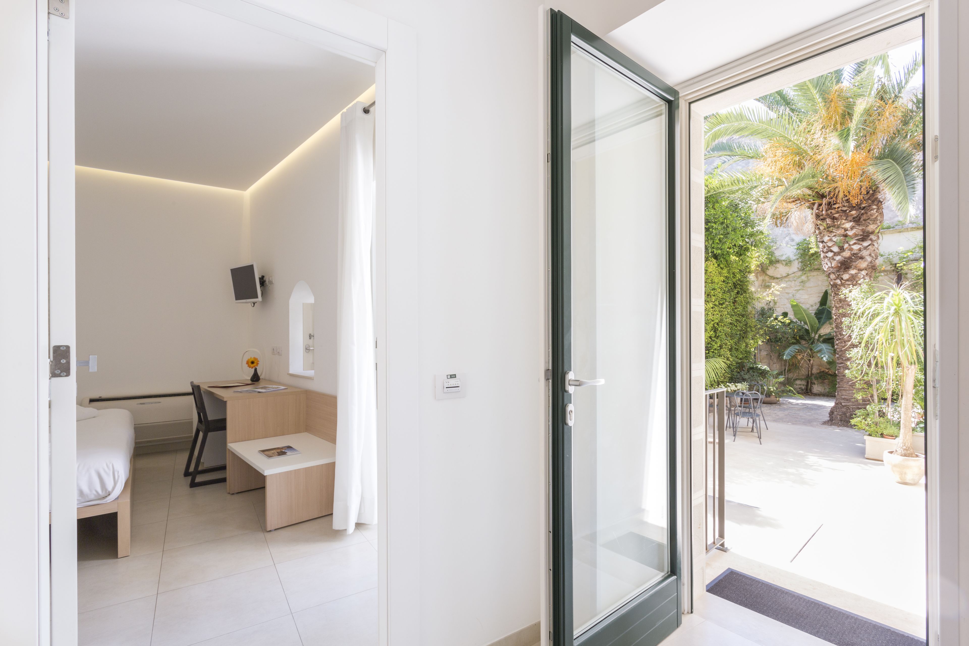 Ein modernes Hotelzimmer mit Bett, Schreibtisch, Stuhl und einer Glastür, die auf einen Balkon mit Pflanzen und einem Palmenbaum führt.