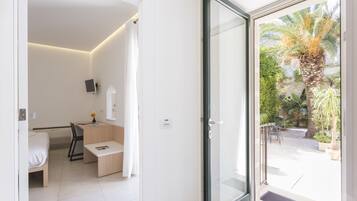 Ein modernes Hotelzimmer mit Bett, Schreibtisch, Stuhl und einer Glastür, die auf einen Balkon mit Pflanzen und einem Palmenbaum führt.