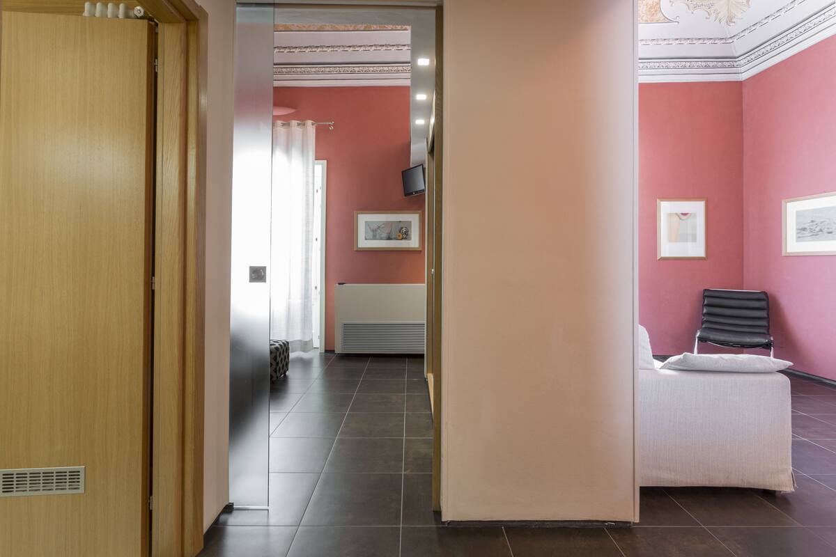 A hotel room with a wooden door, a bed with a white headboard, a black chair, and a pink wall.