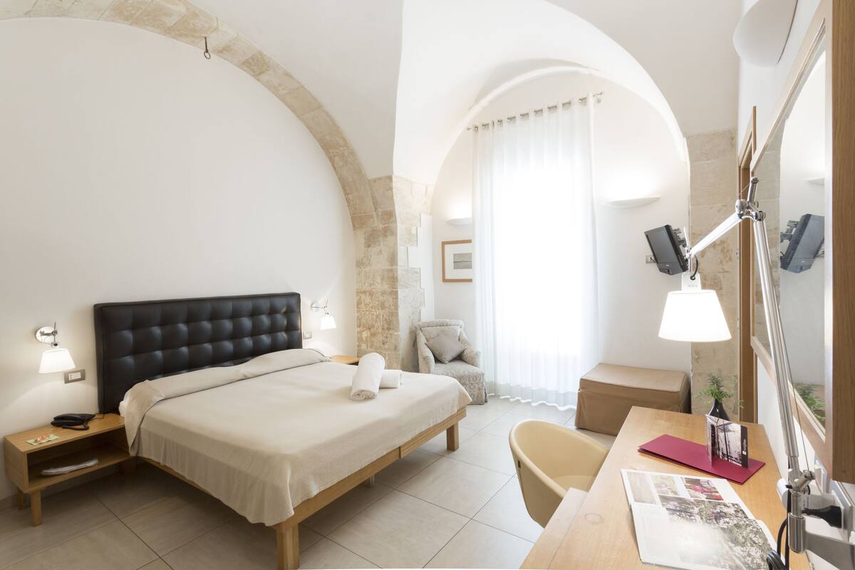 A hotel room with a bed, bedside table, lamp, chair, and a desk with a lamp.