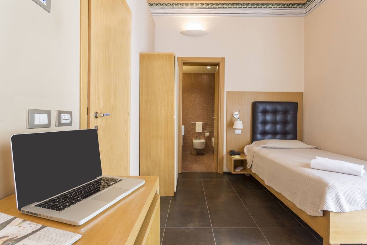 A hotel room with a bed, a desk with a laptop, a bedside table, a bathroom visible through an open door, and a small nightstand with a lamp.