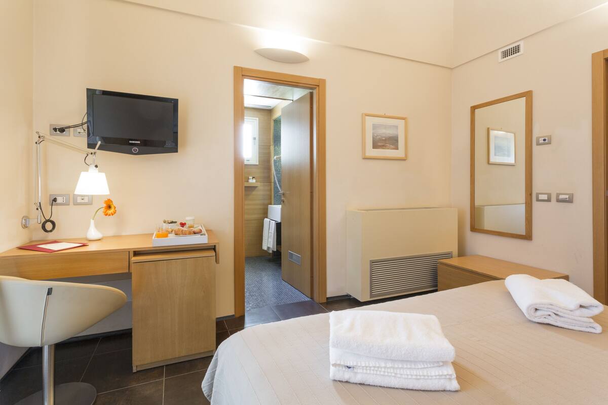 A hotel room with a bed, a desk with a lamp, a TV, and a bathroom visible through an open door.