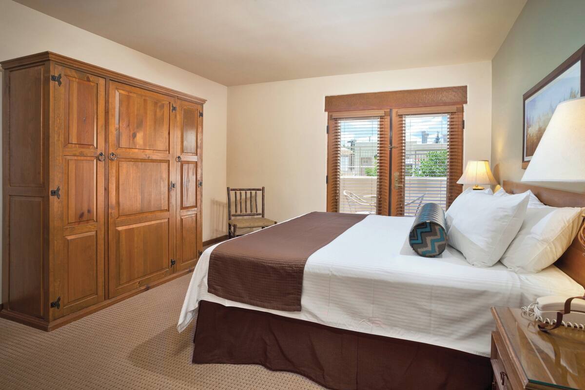 Ein Hotelzimmer mit großem Holzschrank, einem Bett mit weißer und brauner Bettwäsche, einem Nachttisch mit Lampe und einem Fenster mit Jalousien.