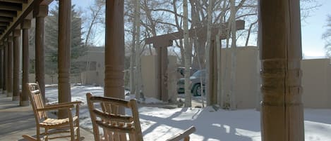 Un porche cubierto al aire libre con mecedoras de madera y suelo de concreto, rodeado de árboles y suelo cubierto de nieve.
