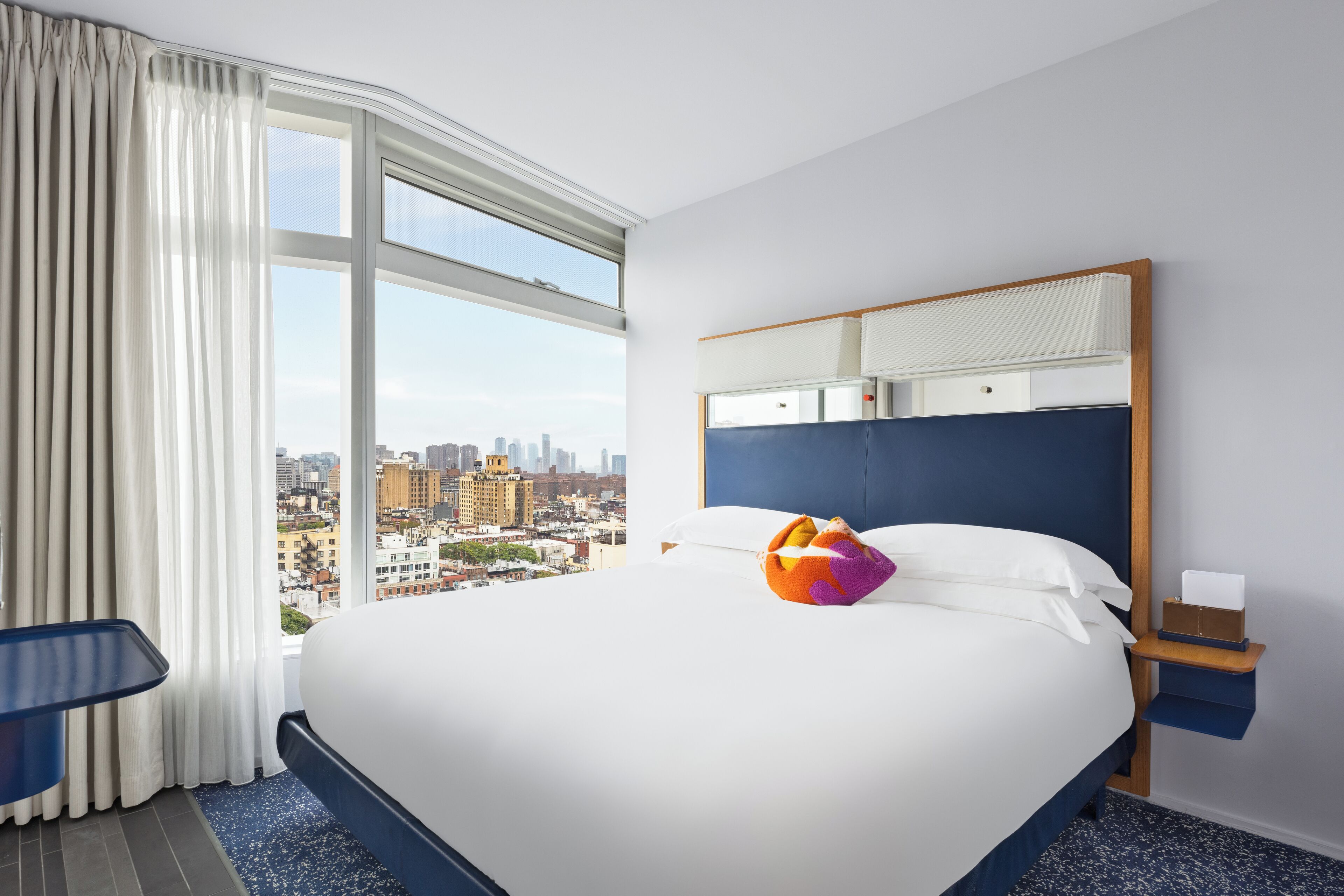 Una habitación de hotel moderna con una cama grande, vistas a la ciudad y una mesita de noche con lámpara.