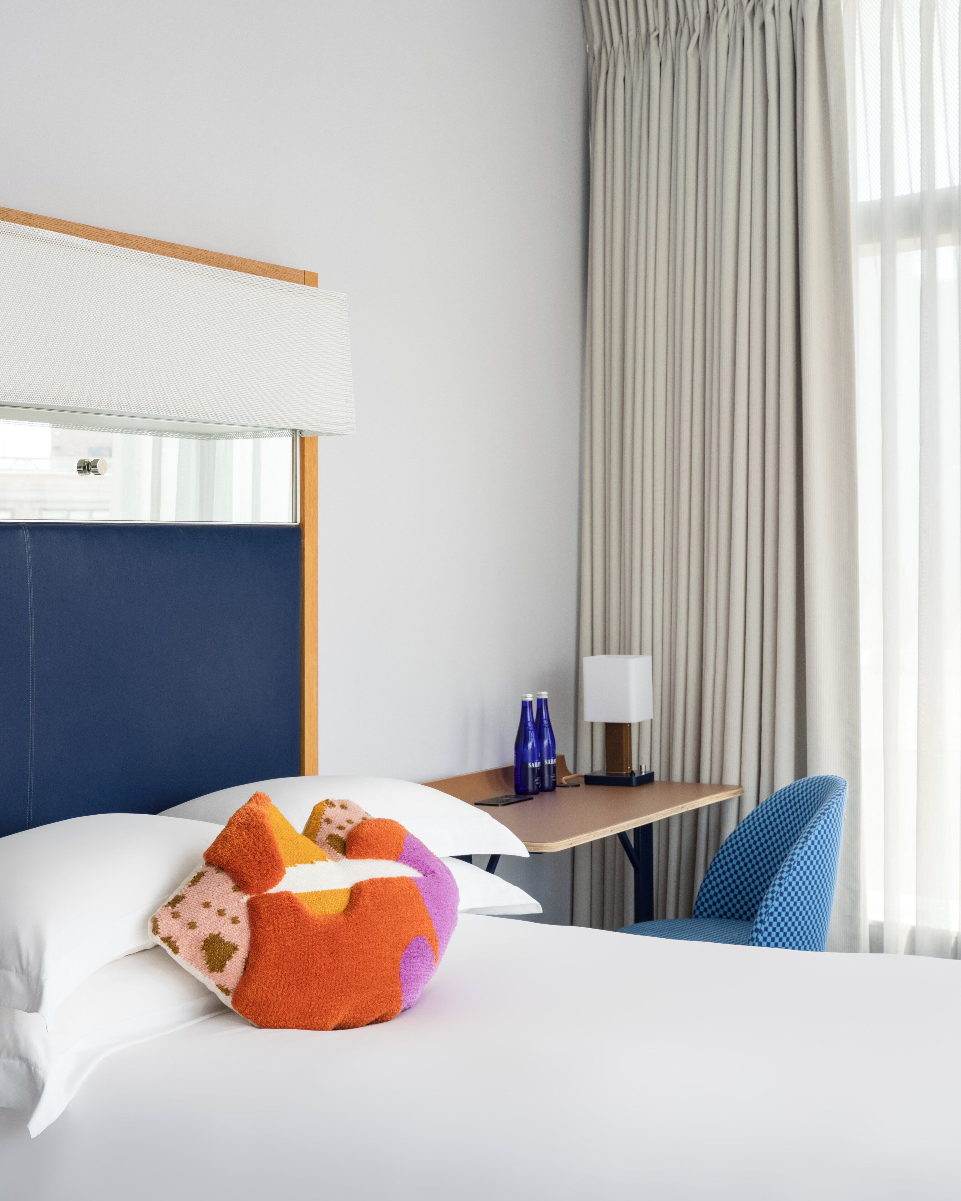 Ett hotellrum med en säng, kuddar, ett skrivbord, en stol, en lampa och gardiner.