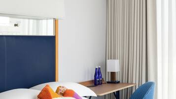 Ett hotellrum med en säng, kuddar, ett skrivbord, en stol, en lampa och gardiner.