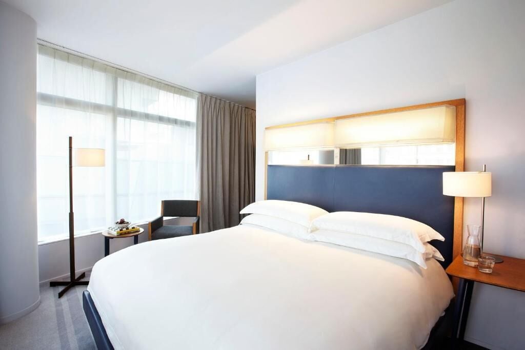 A hotel room with a large bed, a bedside table with a lamp, a chair, a small table with a lamp, and a window with curtains.