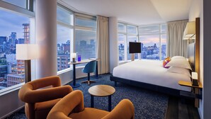 A hotel room with a large bed, two armchairs, a small table, a desk with a chair, a TV, and a city view. - The Standard East Village (New York)