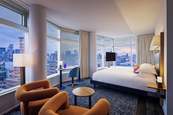 A hotel room with a large window, two orange chairs, a small round table, and a flat-screen TV. at The Standard East Village