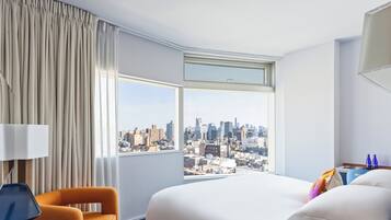 A hotel room with a large bed, a chair, a desk, and a view of a cityscape through a large window.