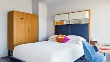 Quarto de hotel com cama, guarda-roupa de madeira, cadeira azul e escrivaninha com abajur.