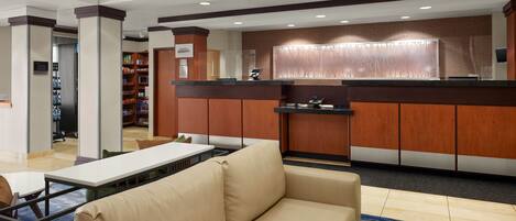 A hotel lobby with a reception desk, a sofa, a coffee table, and a small kitchen area.
