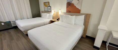 A hotel room with two beds, a bedside table with a lamp, a wall-mounted artwork, and a window with curtains.