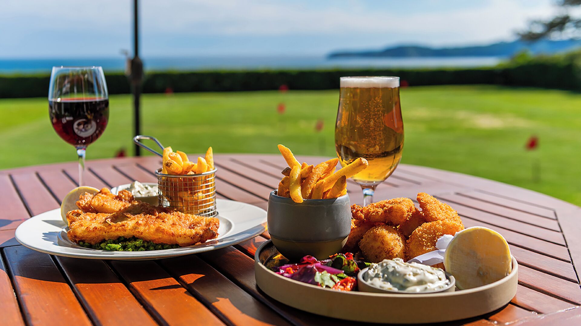 Comida con pescado frito, papas fritas y una cerveza en una mesa de madera, con vistas a un campo verde.