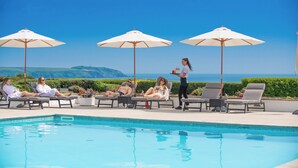 Area piscina con lettini e ombrelloni, una piscina e una vista libera sul mare e le colline sullo sfondo.