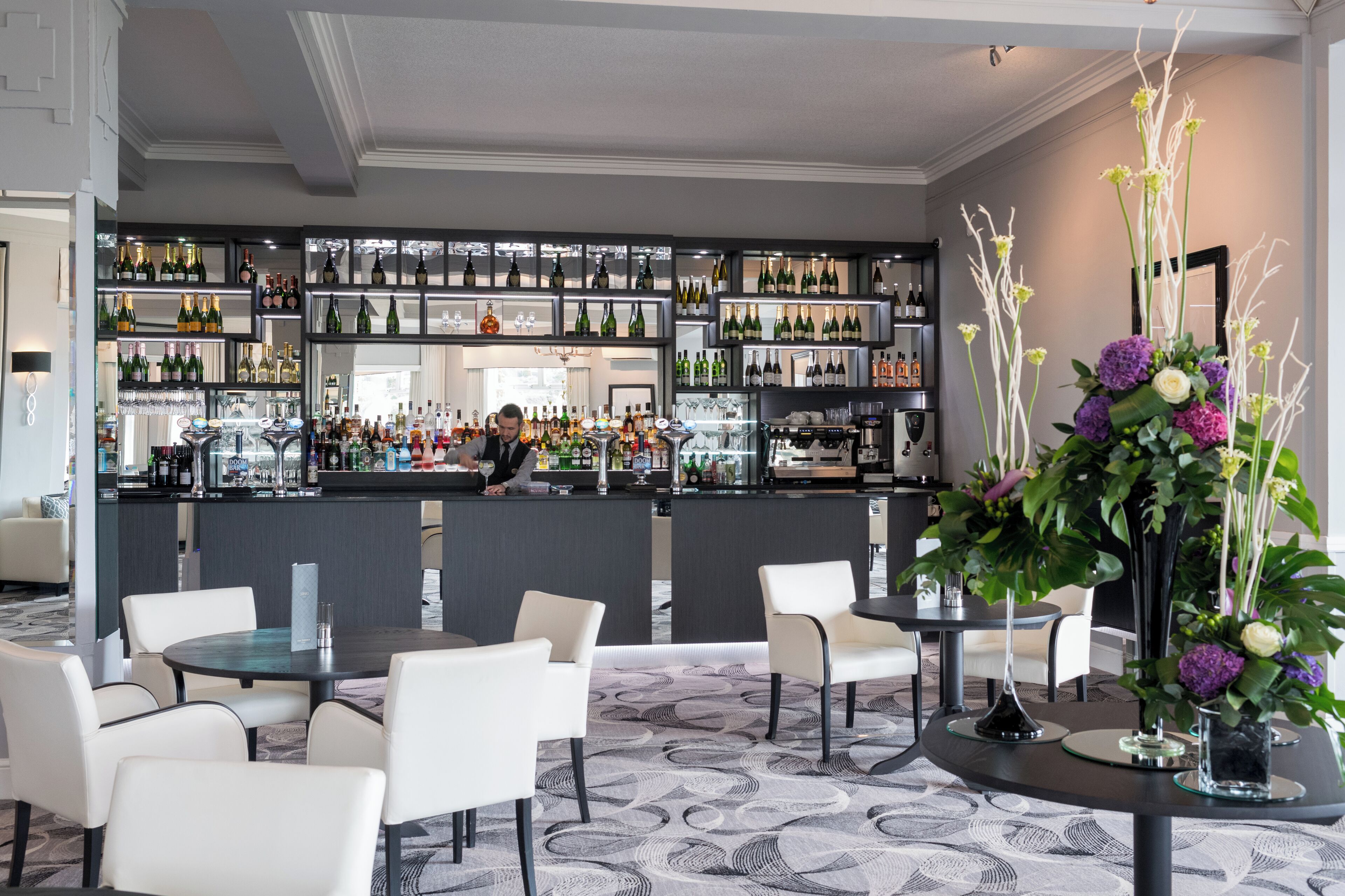 Una zona de bar con varias estanterías llenas de botellas, un barman y diferentes disposiciones de asientos.