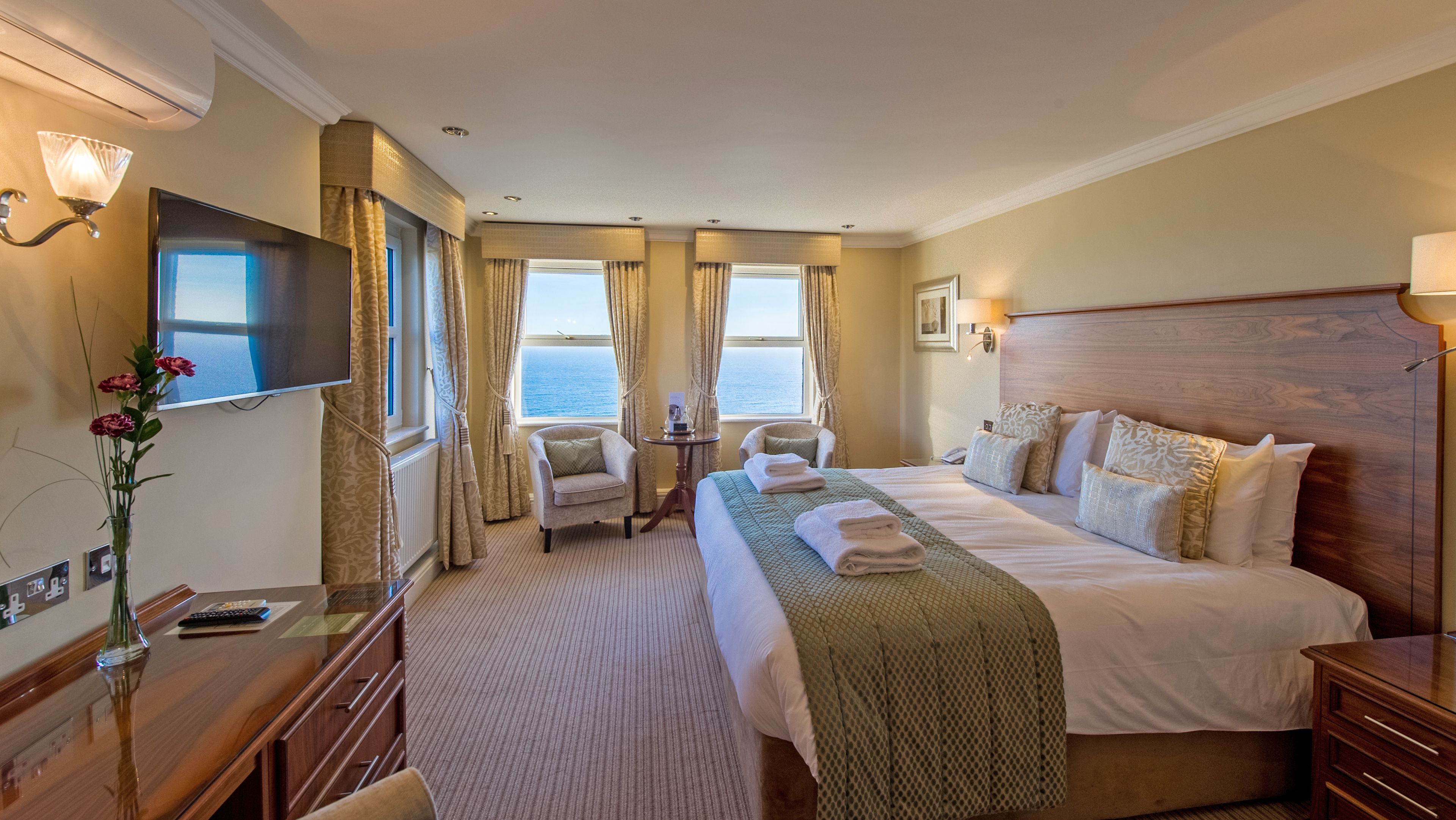 A hotel room with a large bed, two armchairs, a small table, a TV, and a view of the sea.