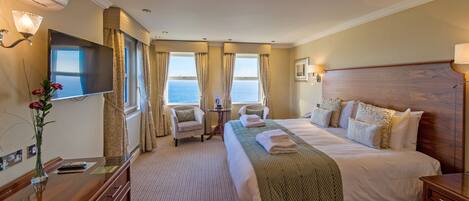 A hotel room with a large bed, two armchairs, a small table, a TV, and a view of the sea.