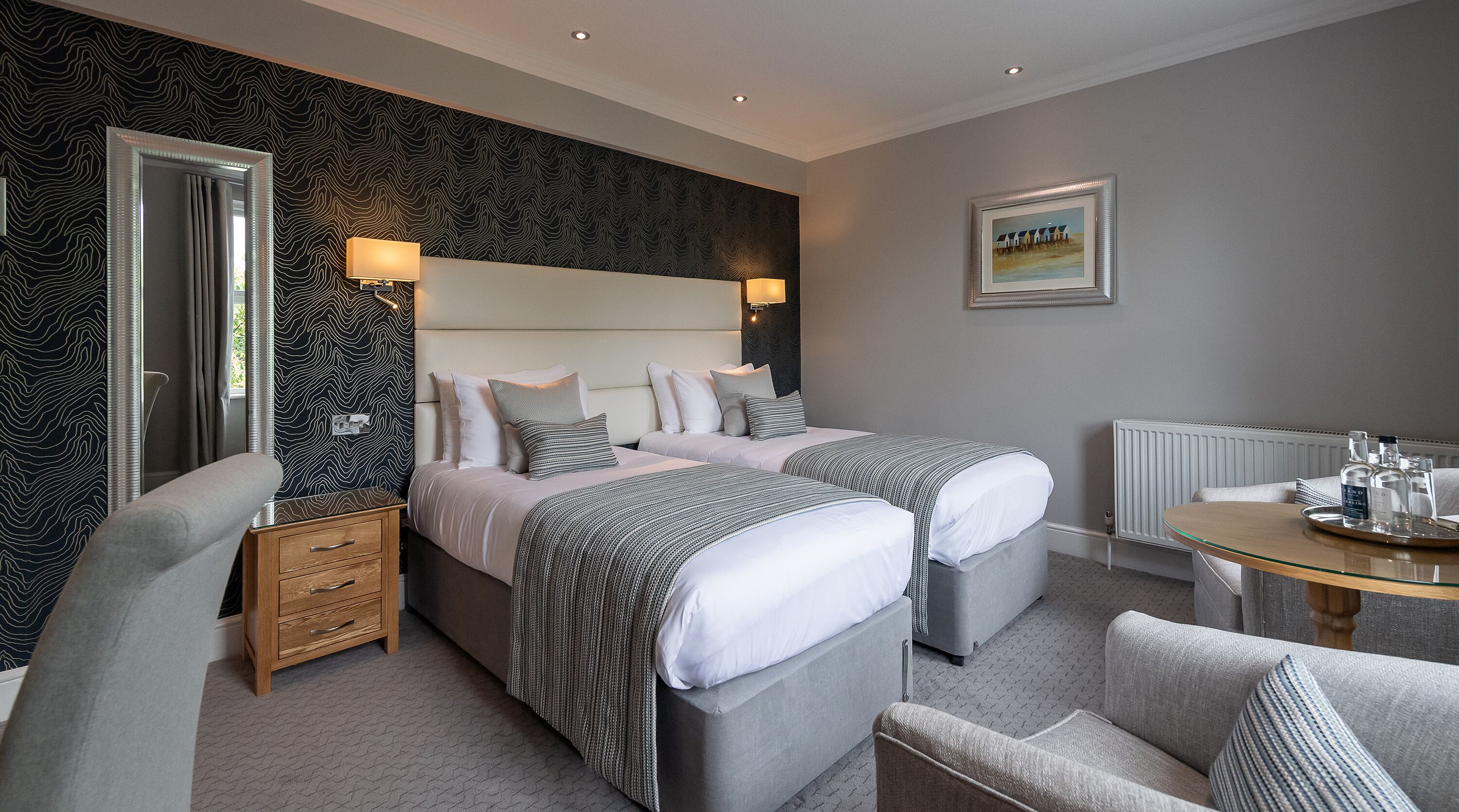 A hotel room with two beds, a wooden nightstand, a chair, a table with a tray, and a painting on the wall.