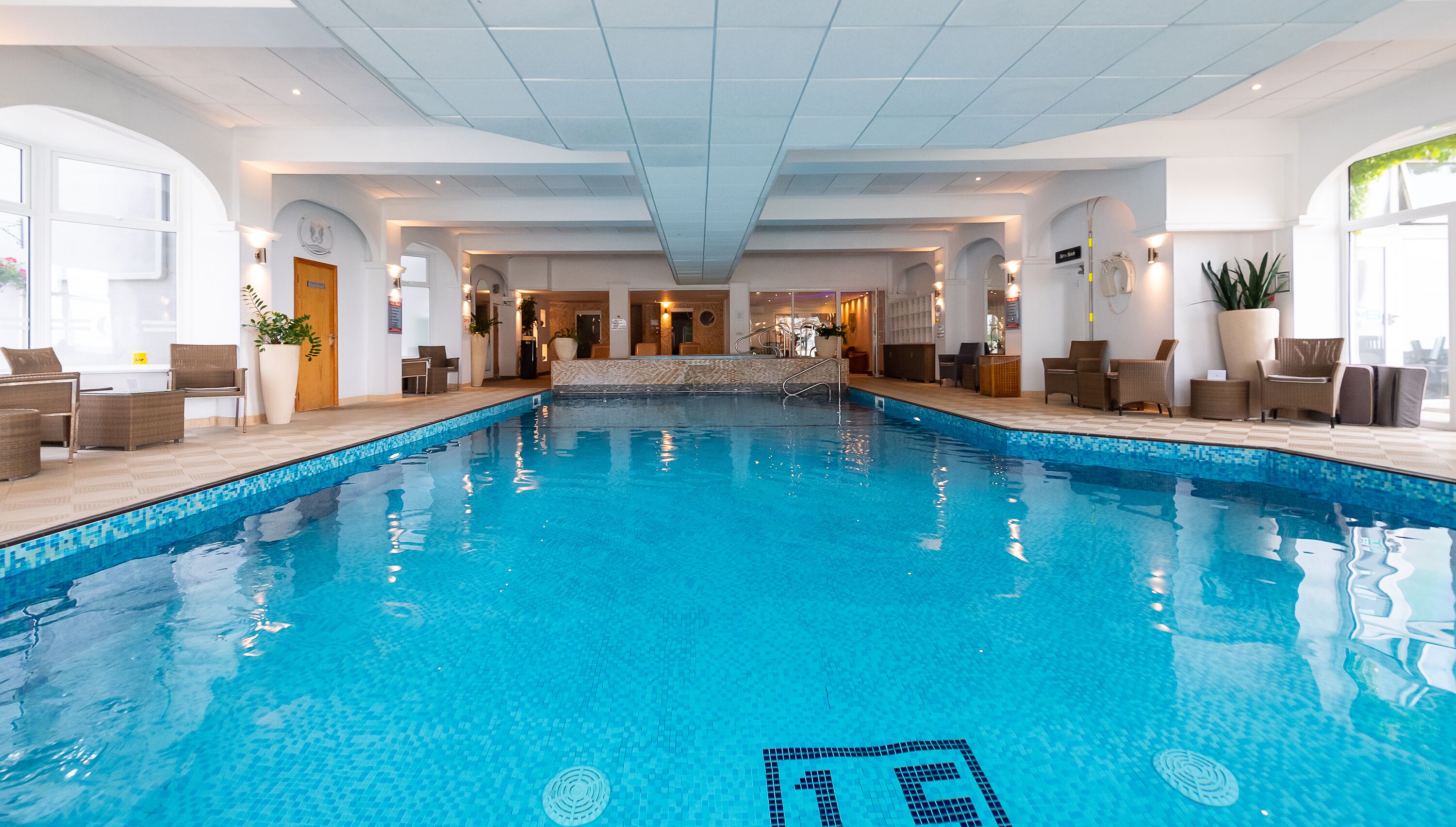 An indoor swimming pool with clear blue water, surrounded by lounge areas with seating and potted plants.