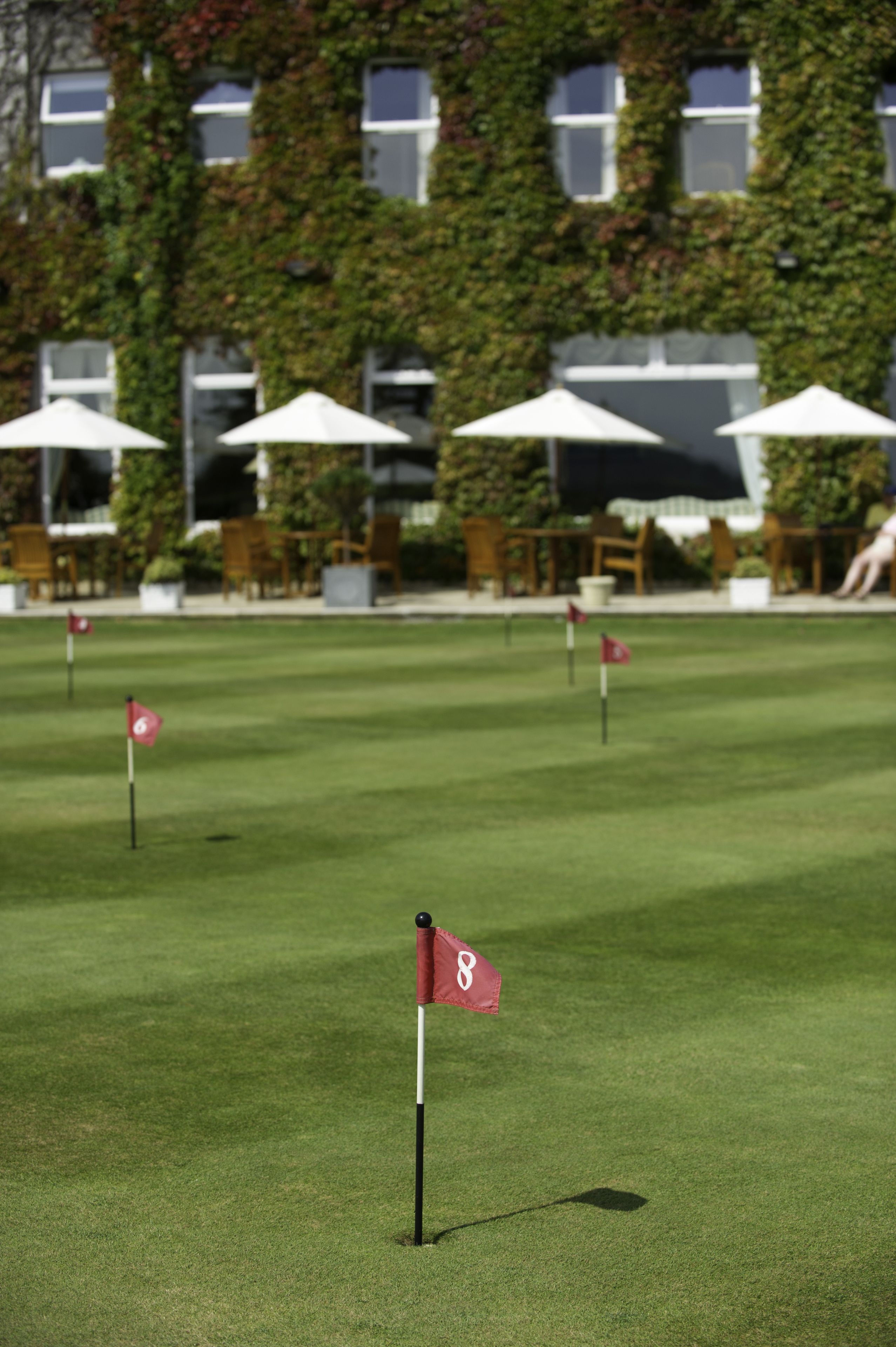 A golf course with a flag marked '8' on the green, surrounded by white umbrellas and a building with ivy-covered walls in the background.