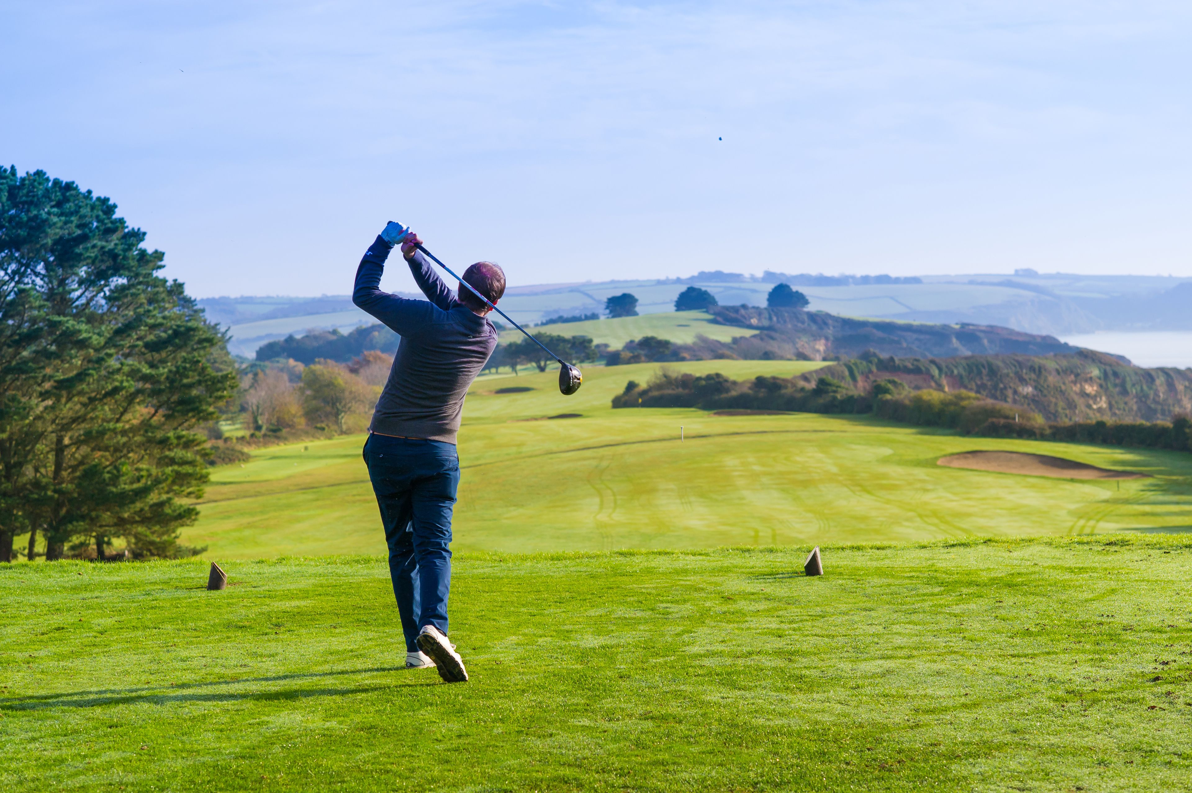 A golfer in mid-swing on a golf course with a scenic view of hills and a body of water in the distance.